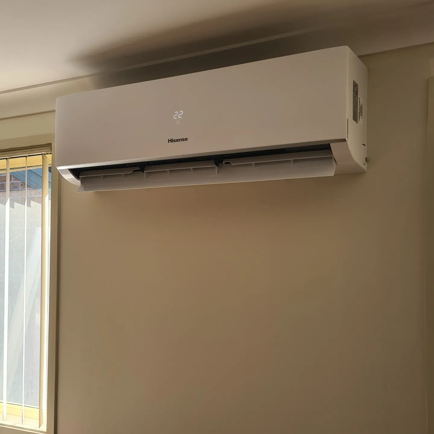 The image shows a modern wall-mounted air conditioning unit in a neutral-toned room. The unit is sleek and white, displaying a digital temperature readout set to 22 degrees Celsius. The design is streamlined, indicating an energy-efficient model. Sunlight filters through a nearby window, highlighting the clean lines of the unit against the wall.