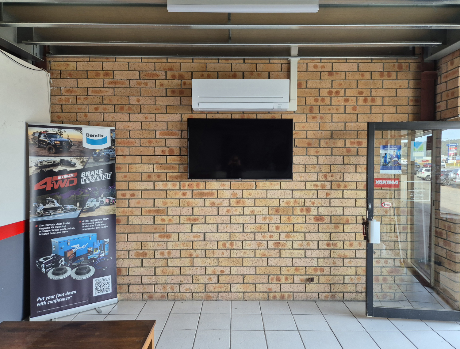 The image depicts a well-lit indoor space with a brick wall featuring an air conditioning unit mounted above a flat-screen television. To the left, there's a promotional banner for auto services, specifically highlighting 4WD brake components. The space appears to be clean and professional, indicating a waiting area or showroom for customers. The layout suggests a customer-focused environment, likely allowing for relaxation while clients await service or consultations. The presence of both an air conditioning unit and electrical services aligns with the Just Chill Air & Electrical business's offerings.