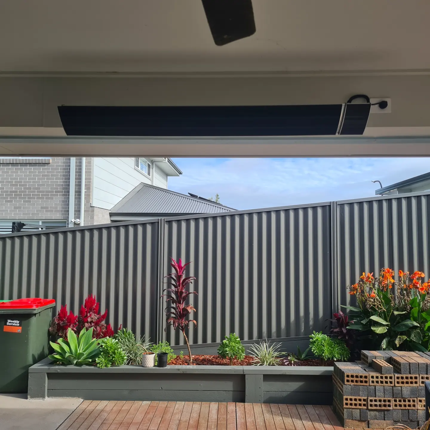 The image shows a covered outdoor area with a wooden deck featuring a variety of plants, including colourful flowers and greenery. A black awning is partially visible above, with a grey fence in the background separating the garden area from a residential building. A green bin is positioned to the left, complementing the vibrant landscape.